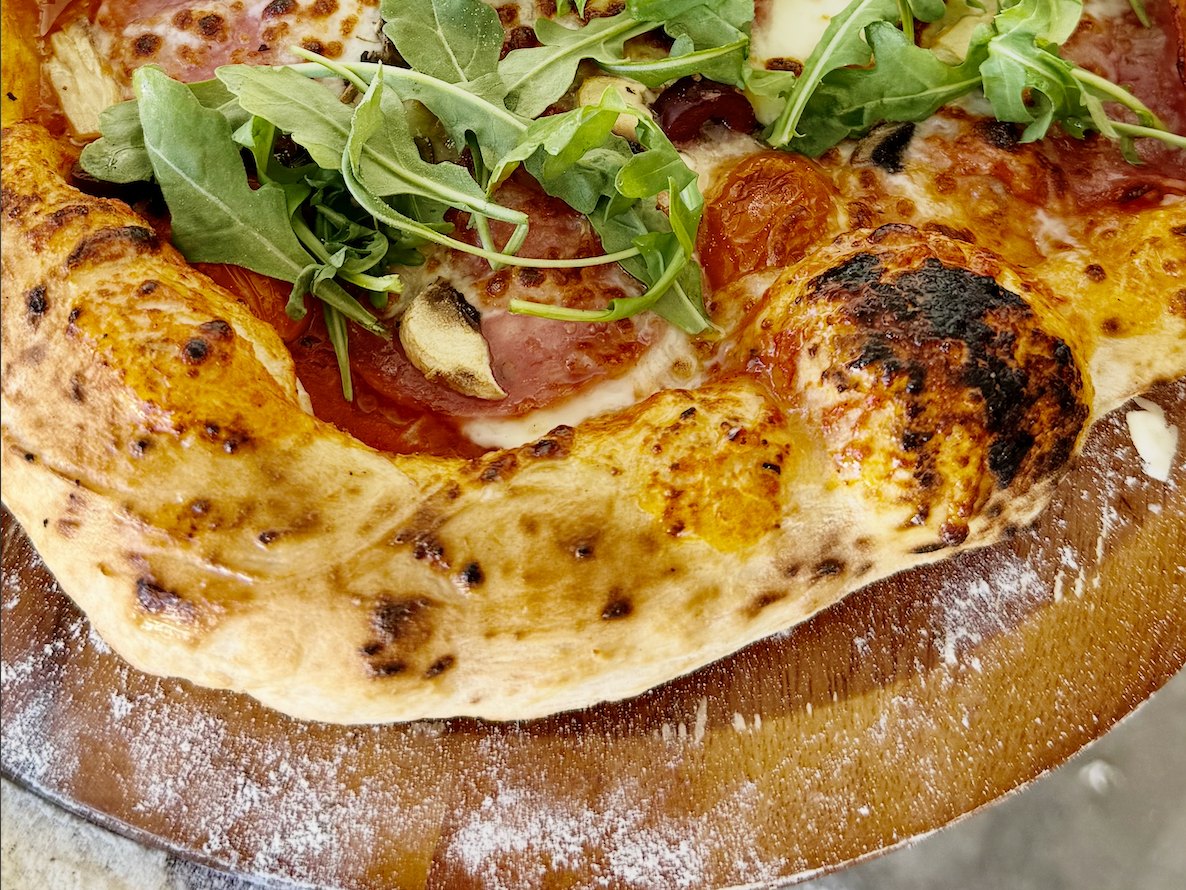 A home-baked pizza with salami, mushrooms and arugula, its puffed cornicione blistered and charred on a floured wooden peel